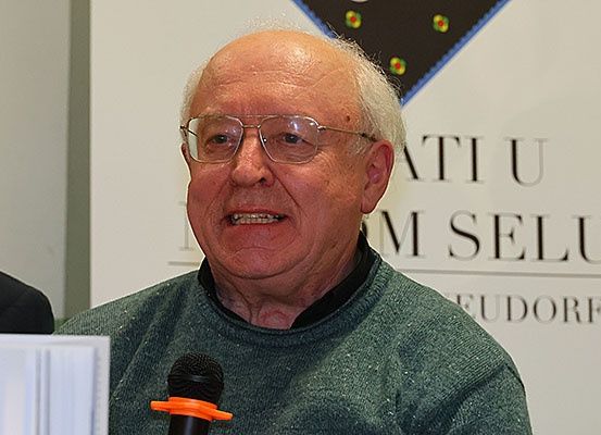 Ein älterer Mann mit Brille spricht in ein Mikrofon. Er trägt einen grünen Pullover und befindet sich wahrscheinlich bei einer Veranstaltung. Hinter ihm befindet sich ein Banner mit dem Text 'IM SELU EUDORF'.