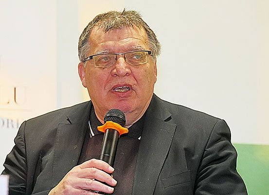 Ein Mann in einem schwarzen Anzug und mit Brille spricht in ein Mikrofon, möglicherweise bei einer Veranstaltung. Hinter ihm ist ein weißer Hintergrund mit verschwommenem Text.