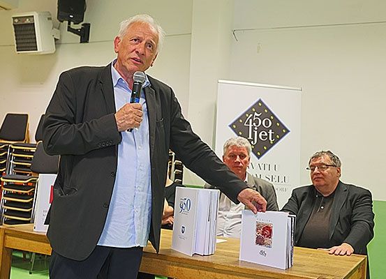 Ein älterer Mann spricht in ein Mikrofon an einem Tisch, mit Büchern und zwei Männern im Hintergrund. Hinter ihm befindet sich eine Wand mit einem Logo und einem Banner.