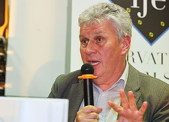Ein Mann mit grauen Haaren spricht in ein Mikrofon auf einer Konferenz. Er trägt eine grüne Jacke und ein weißes Hemd. Er scheint in eine ernsthafte Diskussion vertieft zu sein.