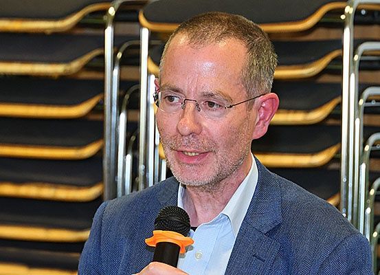 Ein Mann mit Brille, Bart und blauer Jacke hält ein Mikrofon in einem Raum mit vielen gestapelten Stühlen im Hintergrund.