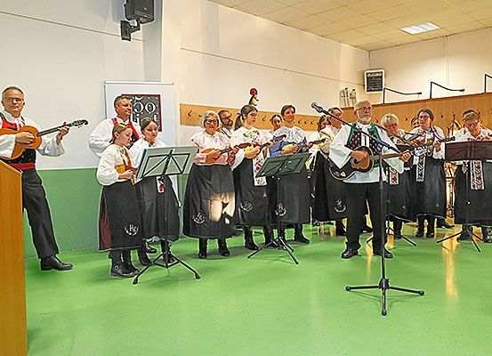 Eine Gruppe von Musikern in traditioneller Kleidung, die verschiedene Instrumente wie Gitarren und Violinen spielen, tritt in einem Raum mit grünem Boden auf.