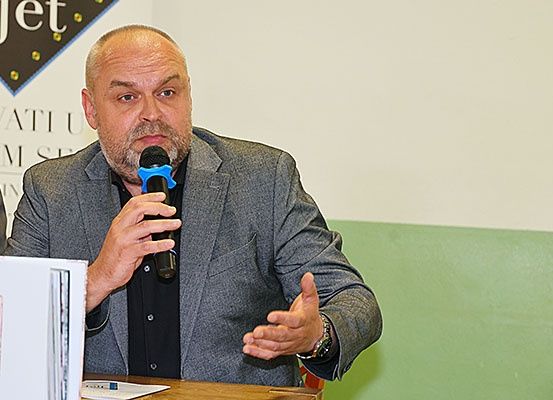 Ein Mann in einem grauen Anzug und schwarzem Hemd spricht in ein Mikrofon auf einer Konferenz und gestikuliert mit den Händen.