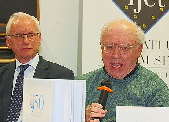 Zwei Männer stehen an einem Podium, einer hält ein Mikrofon und spricht. Hinter ihnen ist ein Hintergrund mit dem Text 'FJET' und einem Logo.