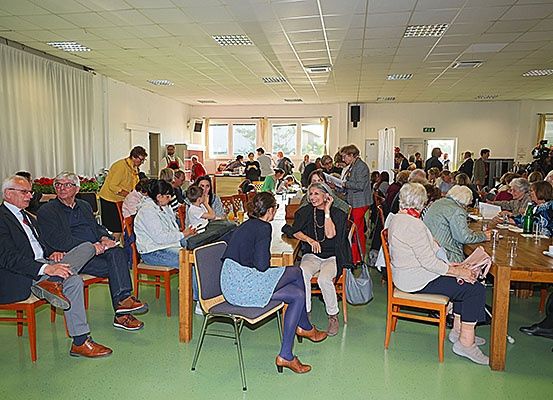 Eine Gruppe von Menschen sitzt auf Stühlen um Tische herum in einem Raum mit grünem Boden, einige lesen Bücher. Im Hintergrund stehen Blumen, und eine Kamera ist vorhanden.