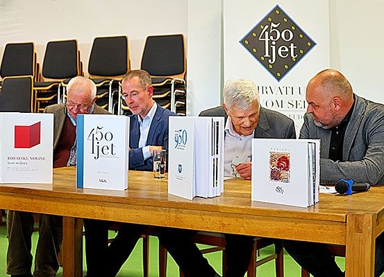 Vier Männer sitzen an einem Tisch mit Büchern. Der Tisch hat eine hölzerne Oberfläche und steht in einem Raum mit Stuhlreihen. Hinter ihnen ist ein Banner mit dem Text 450 ljeto.