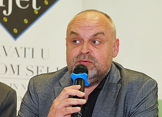 Ein Mann mit Bart hält ein Mikrofon, möglicherweise bei einer Pressekonferenz. Der Hintergrund zeigt ein Banner mit den Texten 'JET' und 'RVATI U OM SEI'.