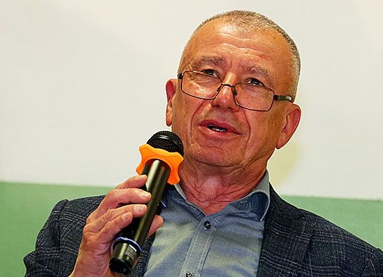 Ein älterer Mann mit Brille und Anzug hält ein Mikrofon mit orangefarbenem Griff. Er steht vor einem grünen und weißen Hintergrund.
