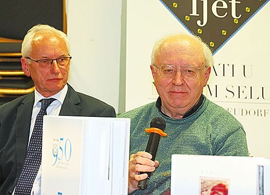 Zwei Männer, einer mit einem Mikrofon in der Hand und sprechend, der andere mit einem Buch, auf dem '950' steht, sitzen vor einer Kulisse mit Text und Logo.