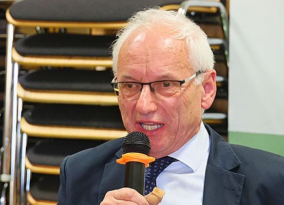 Ein älterer Mann in Anzug und Krawatte hält ein Mikrofon und scheint zu sprechen. Er trägt eine Brille und hat einen Verband an der linken Hand. Hinter ihm sind Reihen von gestapelten Stühlen zu sehen.