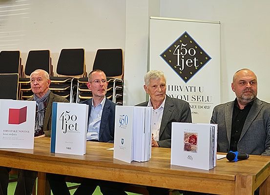 Fünf Männer sitzen an einem Tisch mit Büchern und einem Mikrofon. Dahinter befindet sich ein Schild mit der Aufschrift '450 Jhet'.