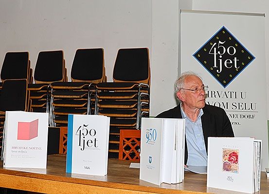 Ein älterer Mann sitzt an einem Tisch mit Büchern mit dem Titel '450 ljet'. Dahinter zeigt eine Hintergrundkulisse den gleichen Titel mit zusätzlichem Text. Gestackte Stühle sind links.