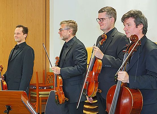 Vier Männer stehen in einem Raum, möglicherweise in einem Musikstudio. Sie sind alle in formeller Kleidung gekleidet und halten Geigen und Celli. Der Mann auf der linken Seite lächelt und der Mann in der Mitte schaut den Mann auf der rechten Seite an.
