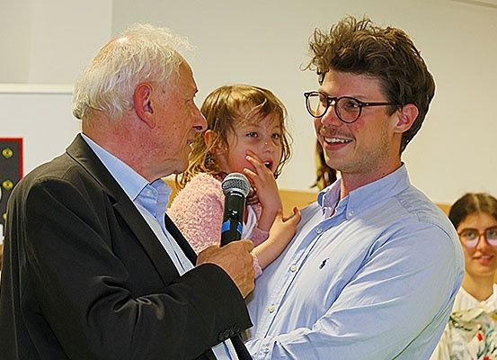 Ein älterer Mann und ein junger Mann halten ein Mikrofon und ein junges Mädchen, möglicherweise bei einer Veranstaltung sprechend. Sie stehen vor einer weißen Wand.