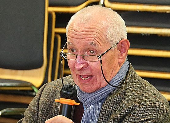 Ein älterer Mann mit Brille und Schal spricht in ein Mikrofon in einem Raum mit Holzstühlen.