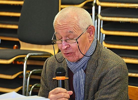 Ein älterer Mann mit Brille und Schal hält ein Mikrofon und scheint in einem Besprechungsraum mit Reihen von Stühlen zu sprechen.