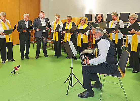 Eine Gruppe Erwachsener, einige mit Brille, führen in einem Raum mit grünem Boden auf, halten Bücher und ein Akkordeon.