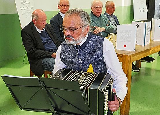 Ein älterer Mann spielt Akkordeon, während er in einem Raum mit anderen sitzt. Hinter ihm steht ein Tisch mit Büchern, auf denen Text und Zahlen stehen.