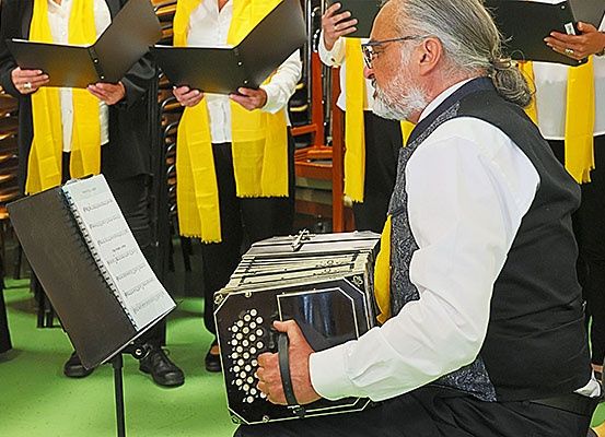 Ein älterer Mann spielt ein Akkordeon, während er mit anderen in einem Chor singt. Hinter ihm halten mehrere Personen Musikblätter. Sie sind in weißen Hemden und gelben Schals gekleidet.