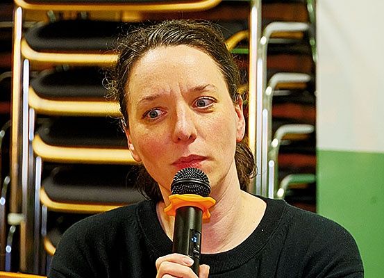 Eine Frau spricht in ein Mikrofon in einem Konferenzraum. Sie trägt ein schwarzes Shirt und kurze Haare. Hinter ihr stehen Reihen von Stühlen.