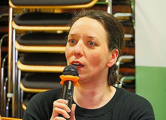 Eine Frau spricht in ein Mikrofon in einem Raum mit Holzstühlen und einer grünen Wand im Hintergrund.