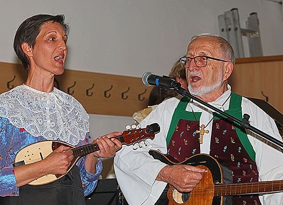 Eine Frau und ein älterer Mann treten auf der Bühne auf, singen und spielen Gitarren. Die Frau hält eine Mandoline, und der Mann trägt eine grüne Schürze mit einem Kreuzanhänger. Sie stehen vor einem Mikrofon.