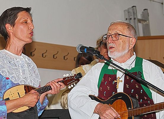 Zwei Personen spielen Gitarren, eine singt in ein Mikrofon, während die andere spielt. Sie tragen beide Langarmshirts und befinden sich in einem Raum mit einer Holzwand.