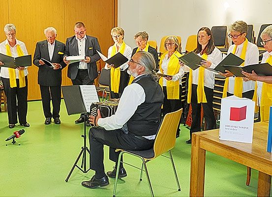 Eine Gruppe von Menschen in gelben Schals singt, während ein Mann ein Akkordeon spielt in einem Raum.