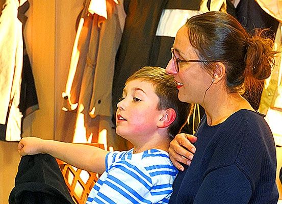 Eine Frau mit Brille spricht mit einem jungen Jungen mit Ohrmarke, beide sitzen, mit Kleidern im Hintergrund.