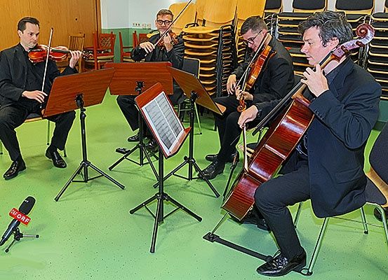 Vier Männer sitzen auf Stühlen und spielen jeweils ein Saiteninstrument. Violinen, Celli und Notenständer sind vorhanden. Ein Mikrofon steht auf dem Boden.
