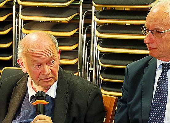 Zwei ältere Männer sitzen nebeneinander in einer Besprechung. Der Mann auf der linken Seite hält ein Mikrofon und scheint zu sprechen. Der Mann auf der rechten Seite trägt eine Brille und hat einen ernsten Gesichtsausdruck. Stapel von Stühlen stehen hinter ihnen.