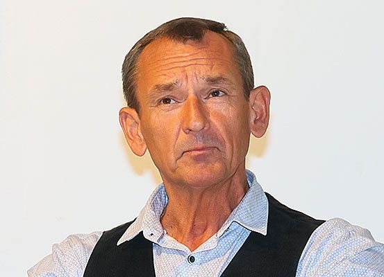 Ein Mann mit ernster Miene, gekleidet in ein blaues Hemd und eine schwarze Weste, scheint zu sprechen oder eine Rede zu halten.