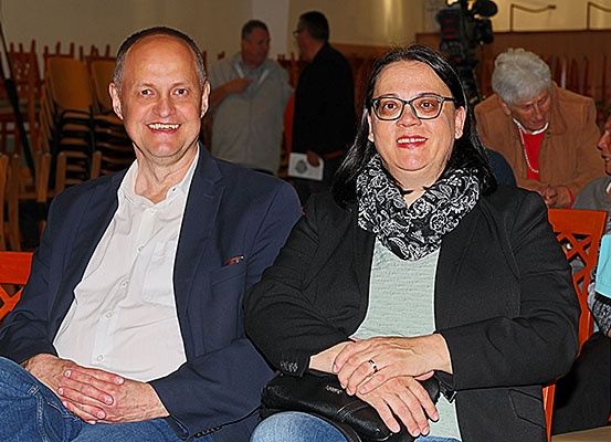 Ein Mann und eine Frau sitzen eng zusammen in einem Raum mit Holzstühlen, lächeln und schauen zur Kamera. Sie tragen eine Brille und einen Ring. Hinter ihnen sitzen Menschen auf Stühlen, einer hält ein Papier.