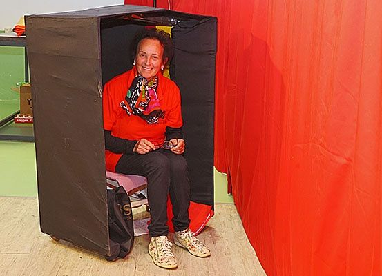 Eine Frau in einem roten Shirt sitzt in einer schwarzen Box mit einem orangefarbenen Vorhang dahinter.