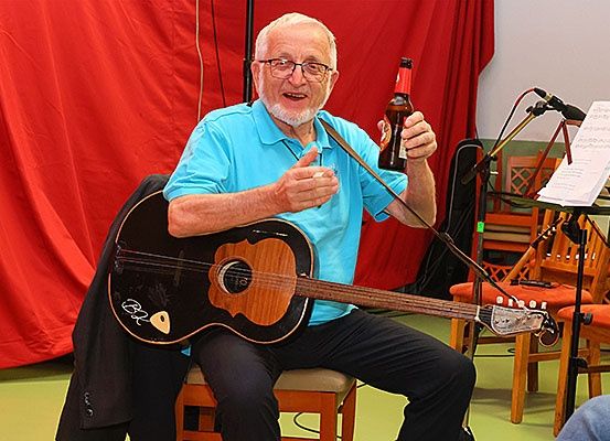 Ein älterer Mann mit Brille und blauem Shirt sitzt auf einem Stuhl, hält eine Gitarre und eine Bierflasche, mit einem roten Vorhang und einem Notenständer dahinter.