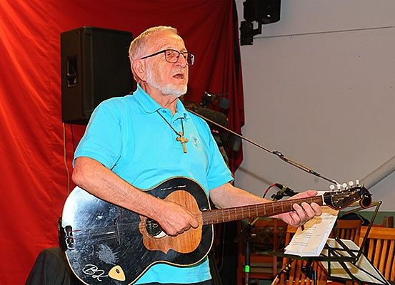 Ein älterer Mann mit Brille und Kreuzkette spielt eine Gitarre und steht vor einem roten Vorhang mit einem schwarzen Lautsprecher.