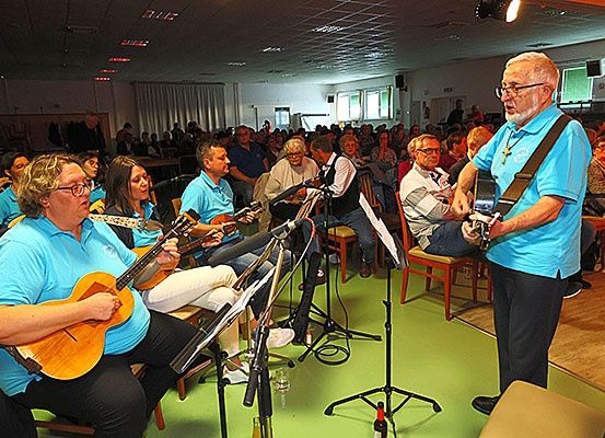 Eine Gruppe von Menschen sitzt auf Stühlen und spielt Gitarren vor einem Publikum, ein Mann steht und spielt Gitarre vor ihnen.