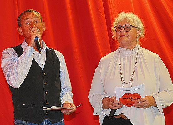 Zwei Personen stehen auf der Bühne; ein Mann spricht in ein Mikrofon, während eine Frau ein Papier mit einem Logo in der Hand hält. Sie stehen vor einem roten Hintergrund.
