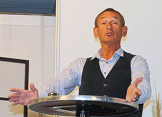 Ein Mann in einer schwarzen Weste steht hinter einem Podium und spricht, während er mit den Händen gestikuliert. Der Hintergrund besteht aus einer weißen Wand und einem gerahmten Bild.