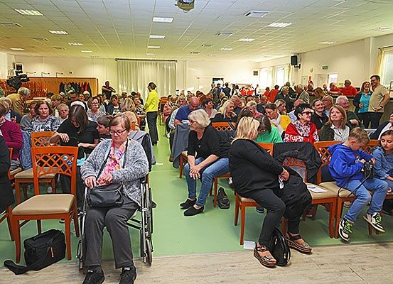 Eine große Gruppe von Menschen sitzt in Stühlen in einem Raum mit grünem Boden. Einige tragen Brillen und Schuhe. Andere tragen Turnschuhe. Eine Frau ist im Rollstuhl.