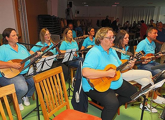 Eine Gruppe von Musikern, alle in blauen Shirts, spielt Gitarren in einem Raum. Einige sitzen, andere stehen. Sie werden von einem Mikrofonständer begleitet. Im Hintergrund sitzen und stehen Menschen.