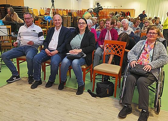 Eine Gruppe von Menschen sitzt auf Stühlen in einem Raum. Einige Leute benutzen ihre Handys. Die Frau im Rollstuhl hält eine Handtasche. Hinter ihnen sitzen Leute auf Stühlen.