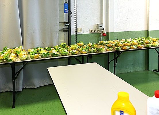 Ein langer Tisch mit vielen Schüsseln Salat darauf, ein weißer Tisch auf der rechten Seite mit einer Flasche Senf darauf, und ein weißer Vorhang im Hintergrund.
