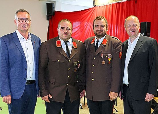 Vier Männer stehen vor einem roten Vorhang, zwei in Militäruniformen und zwei in Anzügen, lächeln für ein Foto.