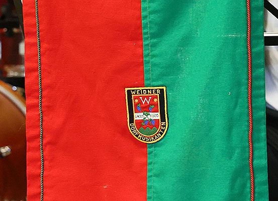 Ein roter und grüner Stoff mit rotem Rand und einem Logo mit der Aufschrift Weidner, D, Dorfmusikanten und LACUS TERRO. Im Hintergrund ist eine Trommel zu sehen.
