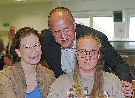 Ein Mann und zwei Frauen sitzen eng beieinander und lächeln in die Kamera. Der Mann trägt einen dunklen Anzug, und die Frauen tragen Freizeitkleidung. Der Hintergrund zeigt einen verschwommenen Raum mit Menschen, einem Tisch und Fenstern.