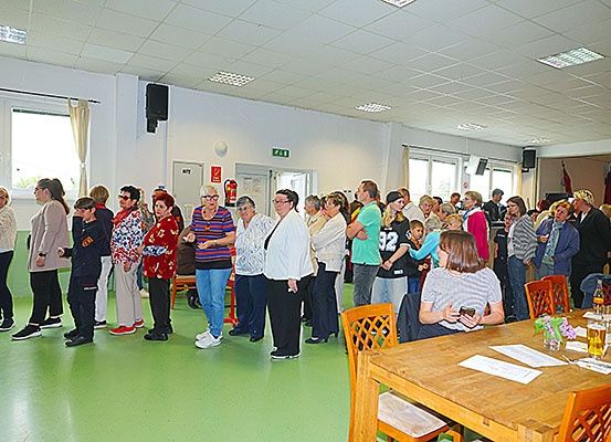 Eine Gruppe von Menschen steht in einem Raum mit grünem Boden und weißen Wänden. Einige tragen Brillen und Turnschuhe. Eine Frau sitzt auf einem Stuhl in der Nähe eines Tisches mit Papieren, Blumen und einem Glas Bier.