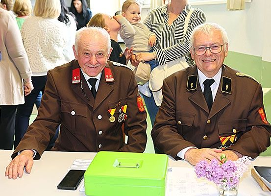Zwei ältere Männer in Militäruniformen sitzen an einem Tisch mit einer grünen Box, einem Handy und Blumen. Einige Leute stehen hinter ihnen.