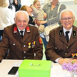 Zwei ältere Männer in Militäruniformen sitzen an einem Tisch mit einer grünen Box, einem Handy und Blumen. Einige Leute stehen hinter ihnen.
