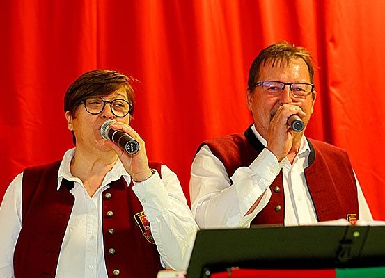 Zwei Personen in roten Westen singen in Mikrofone mit einem roten Vorhang im Hintergrund.